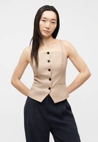 Femme aux longs cheveux noirs portant un haut beige sans manches à boutons et un pantalon noir taille haute, debout les mains derrière le dos.