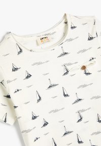 Witte katoenen t-shirt met marineblauwe prints van een zeilboot en een vuurtoren. Ronde halslijn met een kleine houten knop en een touwaccent.