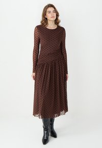 Bruine maxi-jurk met polka-dots in doorschijnende stof, met lange mouwen, getailleerd lijfje, geplooide taille en een vloeiende rok. Gekleed met zwarte laarzen.