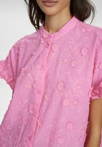 Blusa rosa ricamata con motivi floreali, maniche corte, chiusura a bottoni e tessuto testurizzato.