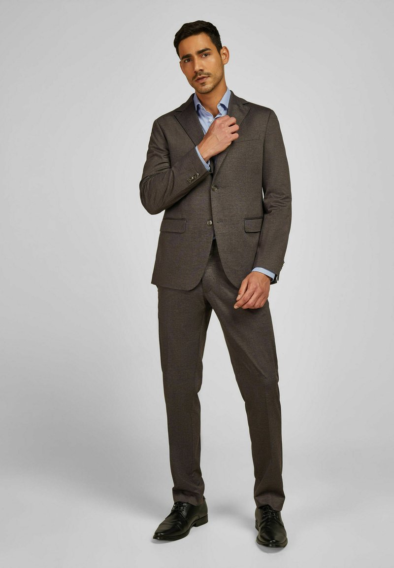 Costume marron sur mesure avec une finition texturée, veste à deux boutons, revers crantés et pantalon à taille plate. Associé à une chemise bleu clair et des chaussures noires.