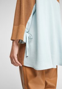 Hnědý pletený top se světle modrou látkou na spodní části, s uvázaným akcentem na boku. Kombinováno s hnědými koženými kalhotami.