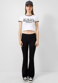 Witte cropped t-shirt met zwarte rand en "Von Dutch Originals" graphic, gecombineerd met zwarte flared leggings en zwarte sneakers met witte zolen.
