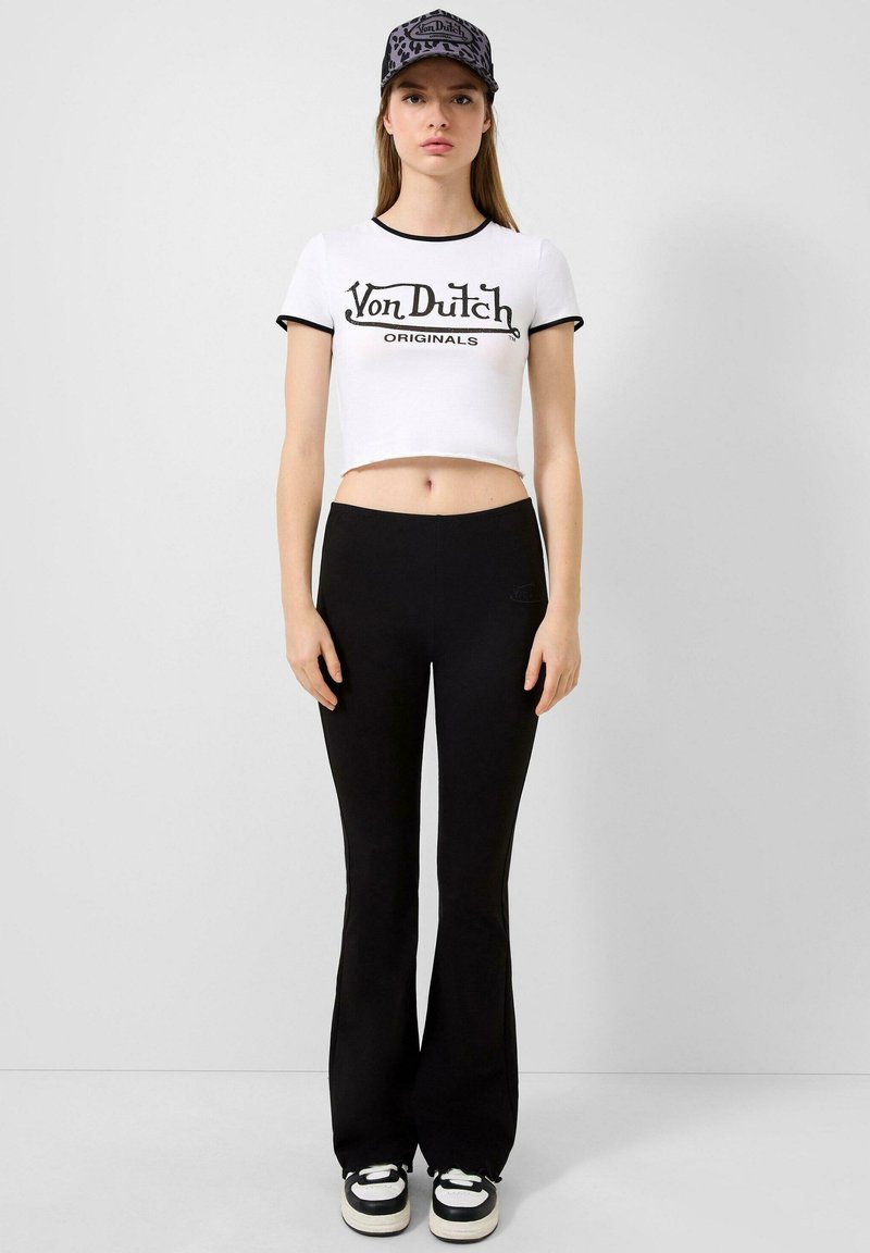 Witte cropped t-shirt met zwarte rand en "Von Dutch Originals" graphic, gecombineerd met zwarte flared leggings en zwarte sneakers met witte zolen.