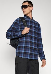 Blue and black plaid flannel shirt with button-down collar, front pockets, worn with dark cargo pants and a black backpack.