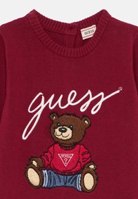 Pull en tricot rouge avec un graphique d'ours brun portant une chemise rouge avec un logo triangulaire, et un texte en cursive blanche "guess" au-dessus.