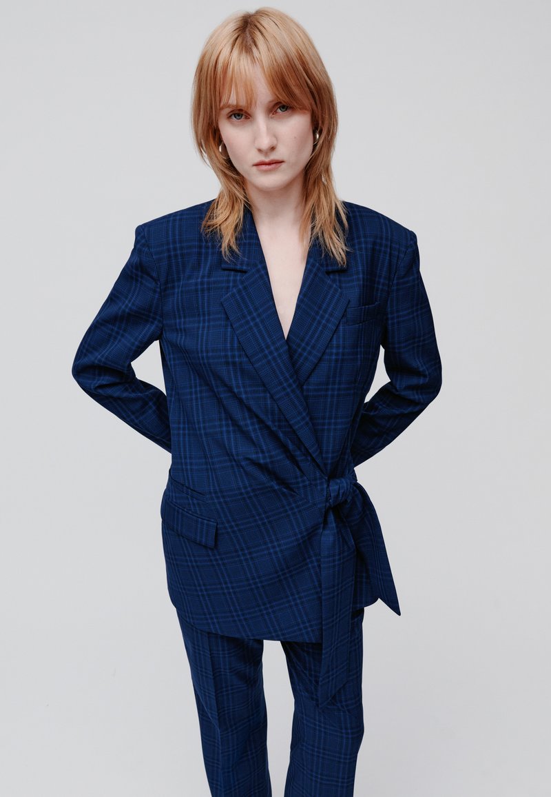 Giovane con capelli rosso chiaro che indossa un completo oversize a quadri blu con vita legata, in piedi con le mani dietro la schiena su uno sfondo chiaro.