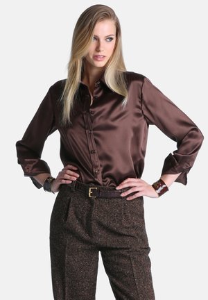 Femme aux longs cheveux blonds portant une chemise en satin marron boutonnée et un pantalon en tweed taille haute avec ceinture, posant les mains sur les hanches.