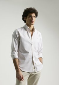 Camisa blanca de botón con collar extendido, mangas largas arremangadas en los puños y un pequeño logo en el pecho. Hecha de un tejido suave.