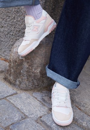 Baskets blanches et rose pâle portées avec des chaussettes blanches et un jean bleu foncé retroussé, un pied reposant sur une surface en pierre rugueuse.