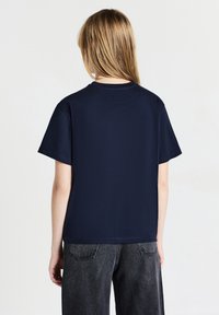 Persona bionda di spalle, che indossa una semplice maglietta a maniche corte blu navy e pantaloni in denim grigio scuro, in piedi davanti a uno sfondo bianco.