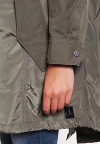 Main reposant sur le côté portant une veste vert olive avec un poignet boutonné et un écusson logo "ck" noir près de l'ourlet, associée à un jean bleu.