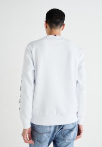Sudadera blanca con cuello redondo y puños acanalados. Presenta una impresión de texto negro a lo largo de las mangas y una etiqueta multicolor en la parte posterior del cuello.