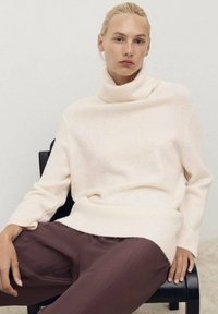 Krema turtleneck pulover iz mehkega, teksturiranega materiala, v kombinaciji z ohlapnimi, temno burgundskimi hlačami. Preprosti, moderni oblikovni elementi.