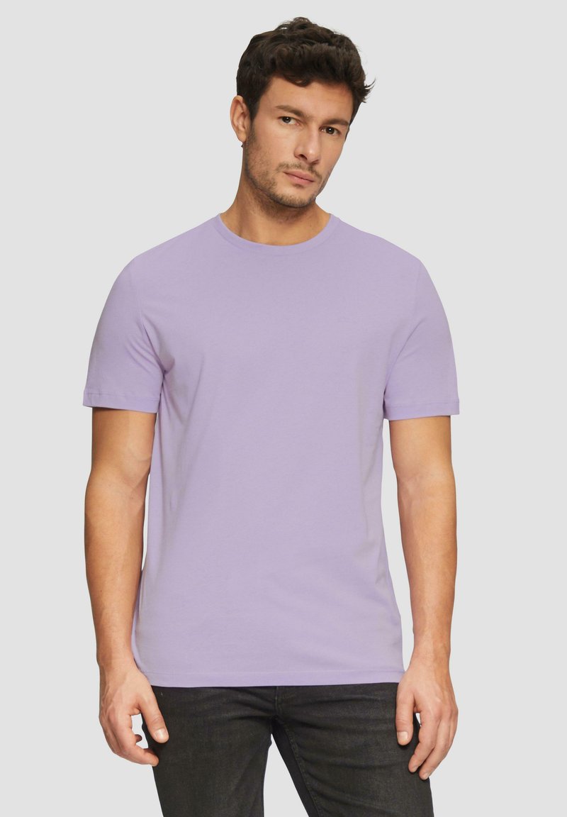 Mężczyzna z krótkimi, kręconymi włosami, wearing a plain lavender short-sleeve t-shirt and black pants, stojący na tle jasnoszarym.