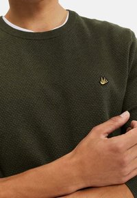 Tmavě zelený strukturovaný svetr s kulatým výstřihem a malým zlatým logem na hrudi. Model má ruce složené před sebou.