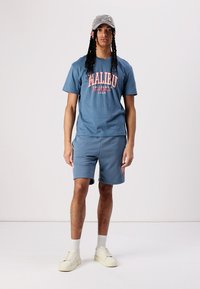 Blauwe katoenen t-shirt met "MALIBU" graphic, gecombineerd met bijpassende blauwe shorts. Model draagt een grijze pet en crèmekleurige sneakers met witte sokken.