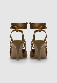 Sandales à talons hauts marron avec un fin stiletto, dos ouvert et brides autour de la cheville, présentant une texture lisse et un éclat subtil.