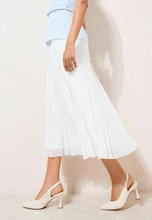 Kvinde gående i hvid plisseret midi-nederdel, lyseblå tekstureret top og cremede slingback-højhælede sko foran en lys væg.