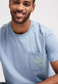 Jasnoniebieska bawełniana koszulka z okrągłym dekoltem, krótkimi rękawami oraz kieszonką z przodu z zielonym graficznym napisem. Gładka tekstura, casualowy design.