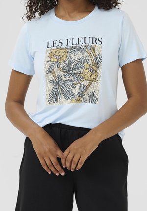 Frau trägt hellblaues T-Shirt mit schwarzem und goldenem floralen Design und dem Text "LES FLEURS" über dem Design.