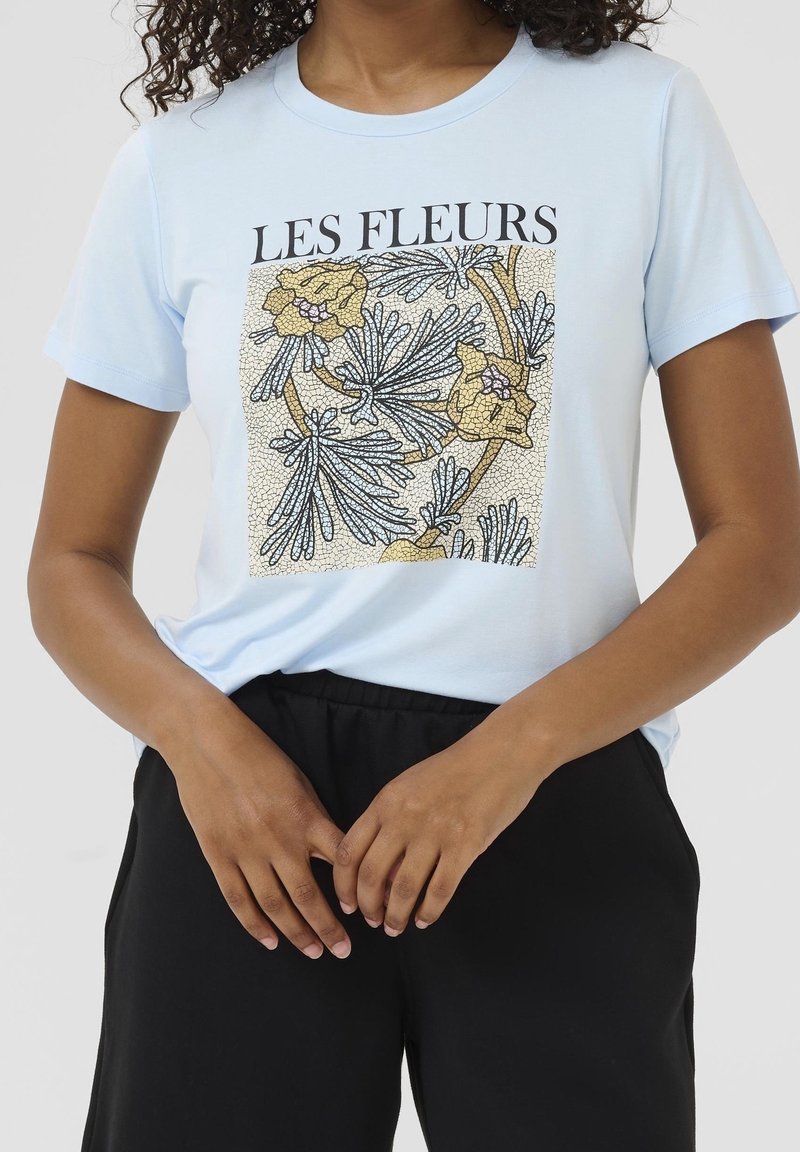 Femme portant un t-shirt bleu clair avec un motif floral noir et or et le texte "LES FLEURS" au-dessus du motif.
