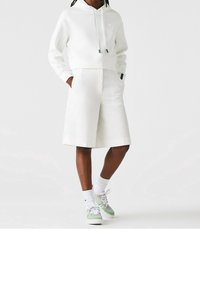Hoodie blanc court avec cordons, associé à un short blanc sur mesure. Chaussures avec des accents verts et chaussettes blanches. Design simple.