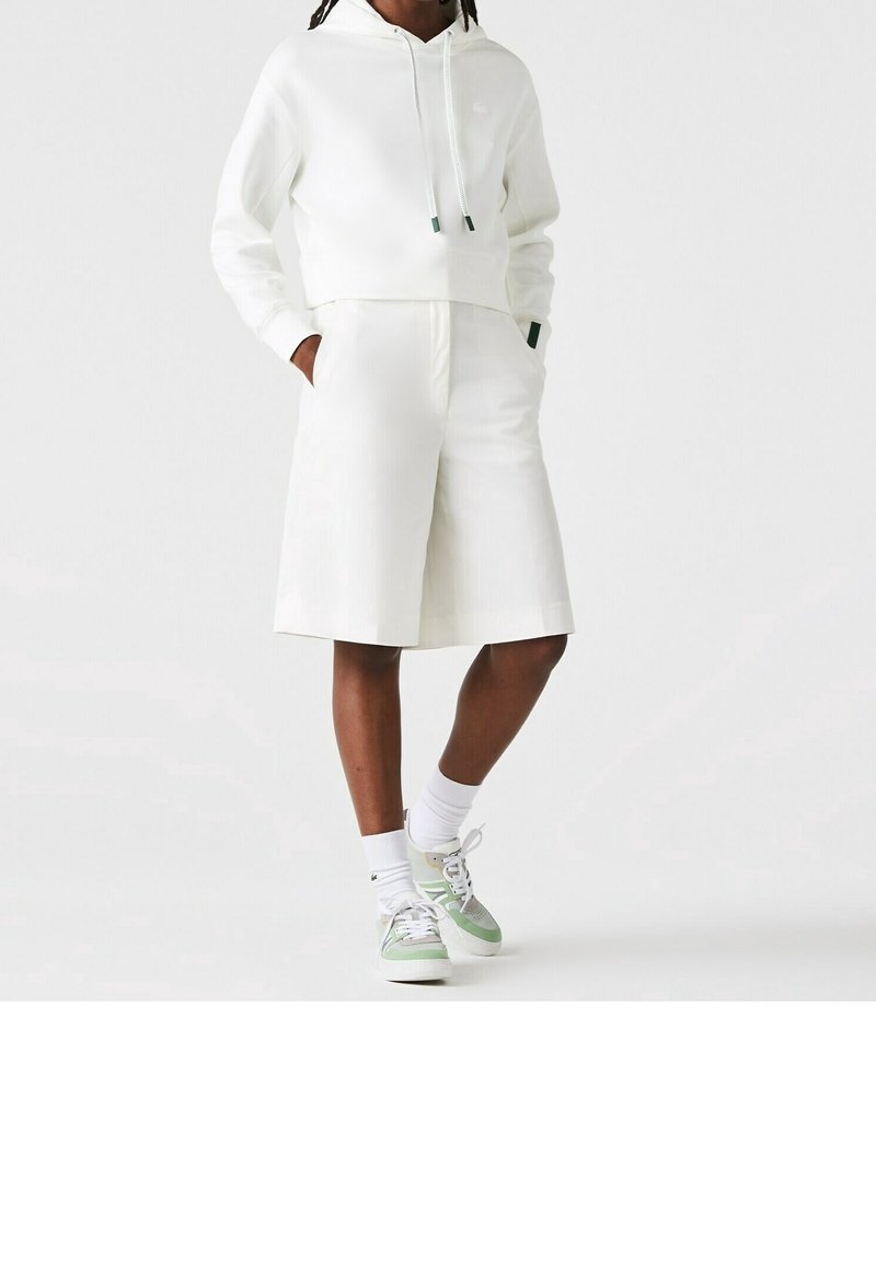Hoodie blanc court avec cordons, associé à un short blanc sur mesure. Chaussures avec des accents verts et chaussettes blanches. Design simple.
