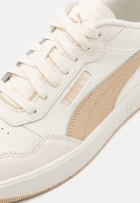 Baskets blanches avec des accents beiges, fabriquées en cuir texturé. Présentent des lacets plats, un détail de logo et une semelle en caoutchouc. Design classique.