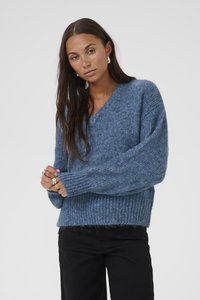 Kék kötött pulóver V-nyakkal, széles ujjakkal és bordás aljjal; puha, bolyhos anyagból készült. Fekete nadrággal párosítva.