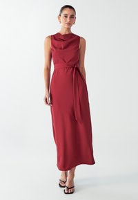 Červené bezrukávové maxi šaty s volným výstřihem a vázáním v pase, vyrobené z hladké tkaniny s lehkým siluetou. Černé sandály na popruzích.