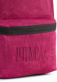 Sac à dos en tissu fuchsia avec fermeture éclair noire, arborant le logo 'PUMA' en relief sur la poche avant. Surface texturée avec finition mate.
