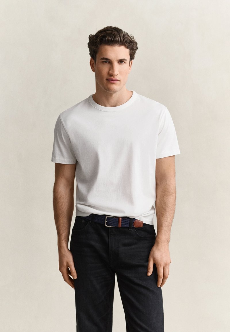 Jeune homme aux cheveux courts et foncés portant un t-shirt blanc uni, un pantalon noir et une ceinture noire avec des détails en cuir marron.