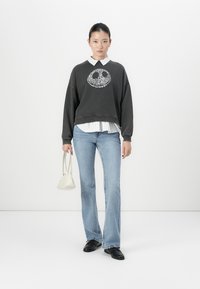 Donna che indossa un felpa nera con un simbolo di pace floreale, una camicia a righe bianche, jeans blu chiaro, scarpe nere, mentre tiene una piccola borsa bianca.