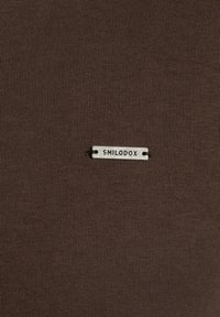 Tela de algodón marrón con una fina textura acanalada. Presenta una etiqueta metálica plateada grabada con "SMILODOX" cerca del centro.