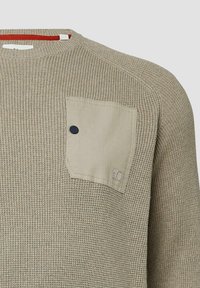 Beżowy sweter dziany z teksturowanym wzorem, okrągłym dekoltem i kontrastową naszywaną kieszenią z ciemnym guzikiem na klatce piersiowej.