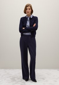 Cardigan navy sopra una camicia a righe bianche e blu, abbinato a pantaloni navy a gamba larga. Il tessuto sembra morbido; i bottoni sono visibili sul cardigan.