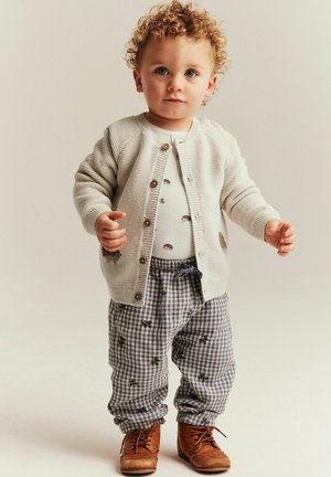 Peuter met krullend haar die een beige vest, een patroonhemd, geruite broek en bruine veterlaarzen draagt, staand tegen een effen achtergrond.