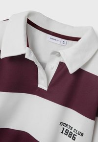 Maroónová a biela pruhovaná polo košeľa s golierom, s gombíkovou légou a potlačou "SPORTS CLUB 1986" v čiernej farbe. Tkanina z bavlnenej zmesi.