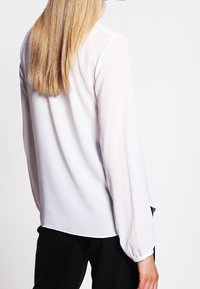 Blusa bianca leggera con maniche lunghe, orlo arrotondato e leggero lucido. Tessuto liscio, senza motivi visibili o dettagli in metallo.