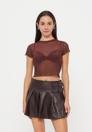 Femme aux longs cheveux bruns porte un top court marron transparent et une mini-jupe plissée en cuir marron foncé taille haute avec des détails à double boucle.