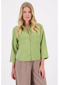 Groene blouse met knopen en lange mouwen, twee borstzakken, zachte stof en een ontspannen pasvorm, gecombineerd met beige wijde broek.