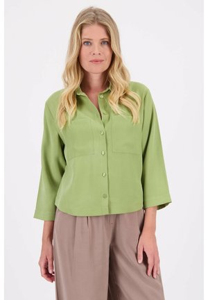 Groene blouse met knopen en lange mouwen, twee borstzakken, zachte stof en een ontspannen pasvorm, gecombineerd met beige wijde broek.
