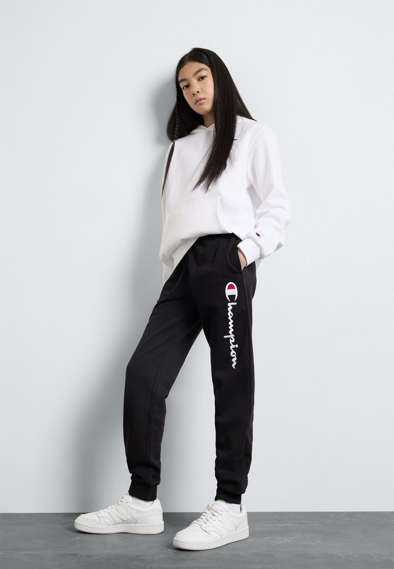 Felpa bianca con cappuccio, pantaloni della tuta neri con logo "Champion" bianco e sneaker bianche. Tessuto morbido, vestibilità rilassata, design minimalista.