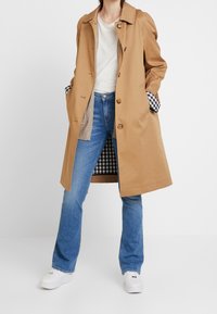 Abrigo beige con un diseño entallado, que presenta un forro a cuadros y cierre de botones. Combinado con jeans acampanados azules y zapatillas blancas.