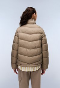 Een bruine pufferjack met een hoge kraag, voorzien van horizontale chevrondetails, een zachte textuur en een losse pasvorm. Draagt over een gestreept T-shirt.