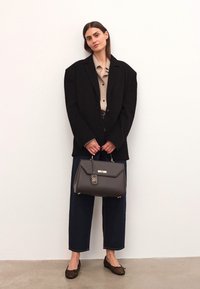 Grå läderhandväska med ett guldaccent och spänne, hålls av en person i en svart överdimensionerad blazer och beige skjorta, kombinerad med vida jeans.