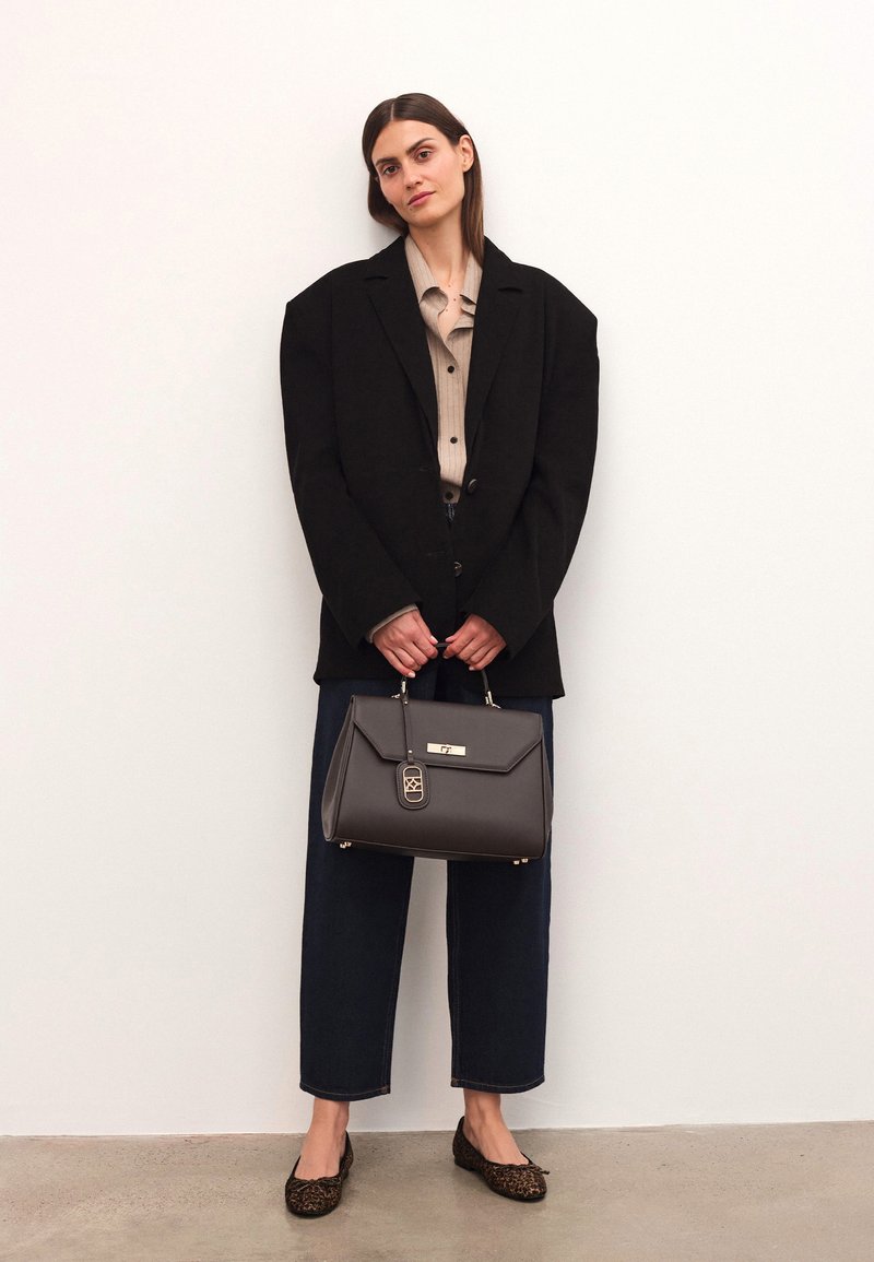 Grå läderhandväska med ett guldaccent och spänne, hålls av en person i en svart överdimensionerad blazer och beige skjorta, kombinerad med vida jeans.