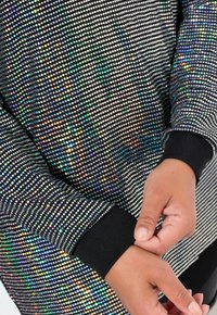 Zwarte sweatshirt met een patroon van iriserende strass-steentjes, textiel met textuur en zwarte ribbelboorden. Handen passen de mouw aan.