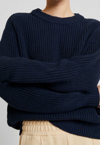 Maglione lavorato a maglia blu navy con texture a coste, maniche ampie e scollatura rotonda, indossato sopra pantaloni beige chiaro. Dettagli includono vestibilità ampia e spalle scese.